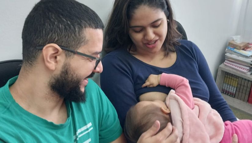 Fotografia de uma mulher e um homem sentados lado a lado. Ela amamenta seu bebê.