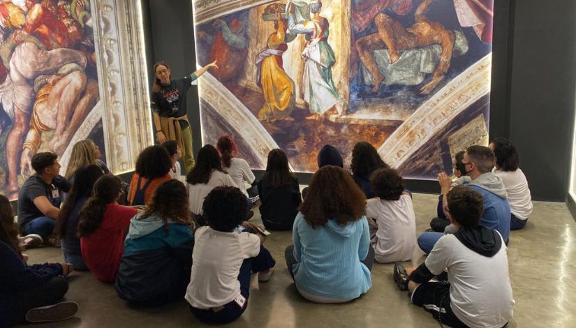 Jovens sentados no chão apreciando uma pintura da exposição "Michelangelo: o Mestre da Capela Sistina", no Museu da Imagem e do Som (MIS) Experience.