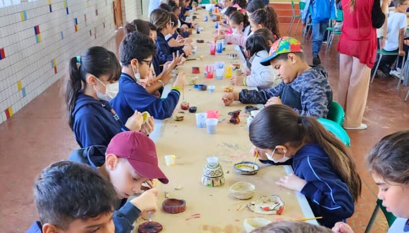 Fotografia mostra estudantes sentados em torno de uma mesa. Sobre a mesa há itens de pintura como tintas e pincéis, eles estão pintando objetos feitos em argila.