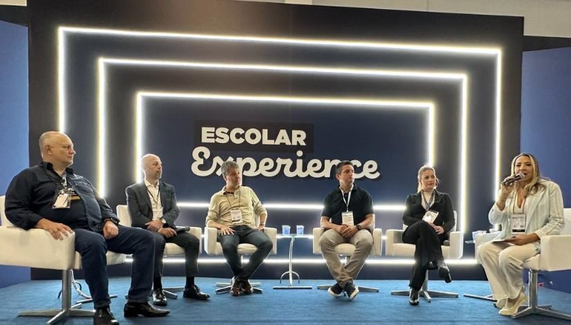 Seis pessoas estão sentadas em cadeiras brancas que estão posicionadas em meia lua. Ao fundo, no telão, está escrito "Escolar Experience".