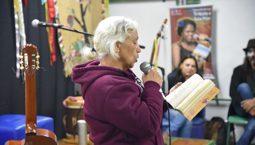 Fotografia de uma mulher realizando a leitura de um livro. Ela segura o microfone e veste uma blusa roxa. Atrás dela, no canto esquerdo da imagem, há um violão. Na lateral direita, em segundo plano, uma mulher a observa e também está aparecendo metade do corpo de um homem com parte do rosto pintado de branco.