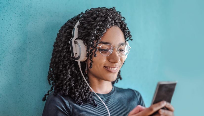 Fotografia de uma mulher sorrindo com fones de ouvidos, óculos e um celular na mão.