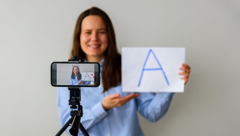 fotografia de uma mulher apresentando um cartaz com a letra A e sendo gravada por uma câmera de filmagem