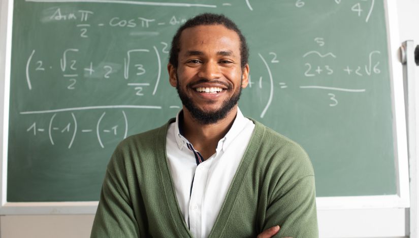 Foto de um homem negro sorrindo de braços cruzados na frente de uma lousa com equações matemáticas.