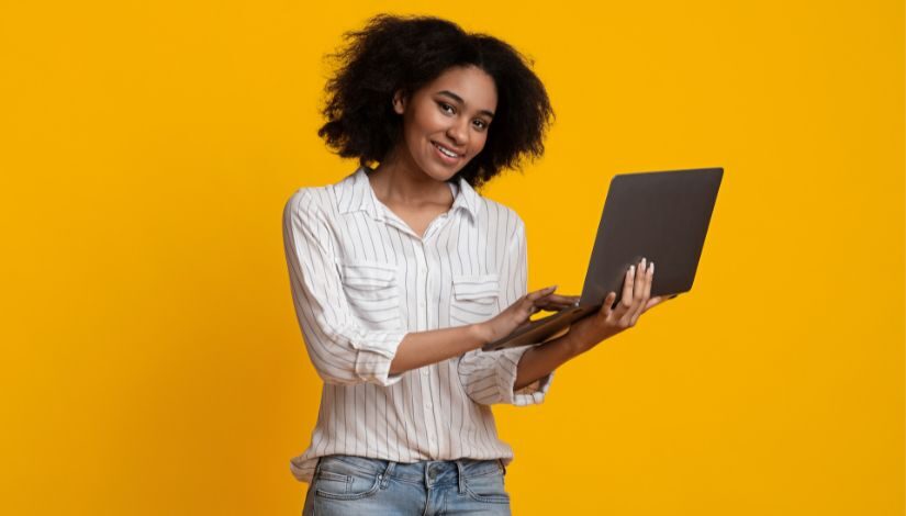 Fotografia com fundo amarelo e, ao centro, uma mulher em pé vestida com camisa branca e calça jeans segura um computador portátil.