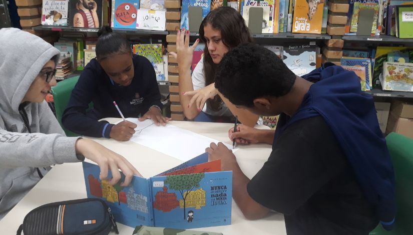 Estudantes estão sentados em torno de uma mesa. Um deles segura um livro de capa azul, outros dois estão escrevendo em um papel e a outra está gesticulando.