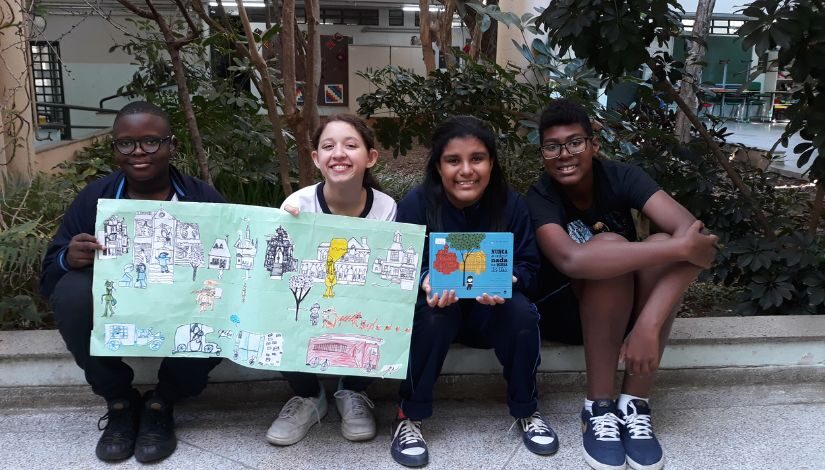 Quatro estudantes estão sentados no chão, Os dois da esquerda estão segurando um cartaz verde com figuras de casas e carros. A terceira menina está segurando um livro com a capa azul e o menino da direita está sentado abraçando as pernas.