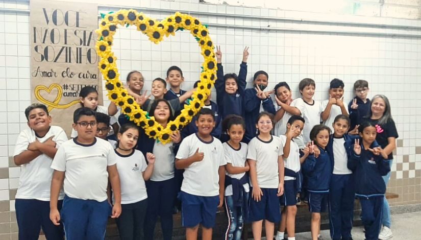 Fotografia mostra grupo de estudantes junto com um painel de girassóis posicionados formando um coração.