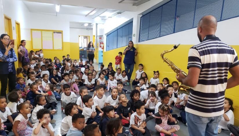 Crianças estão sentadas no chão, elas assistem um homem que está de pé na frente delas tocando saxofone.