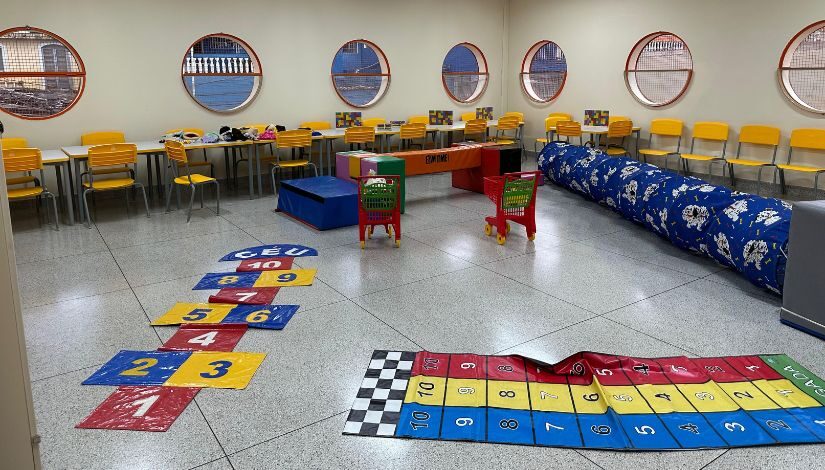 Foto mostra sala com tapetões de brinquedo como amarelinha, corrida dos números. Há também carrinhos de supermercado, túnel de tecido e brinquedos de estimulação e obstáculos. 