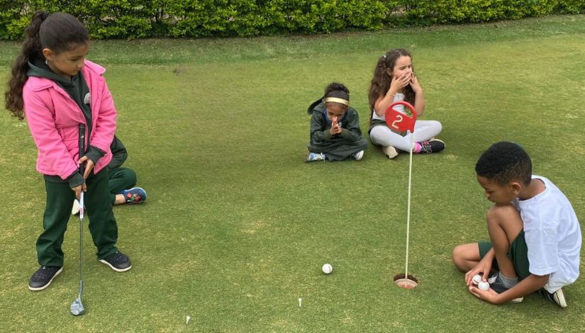 Uma menina está de pé segurando um taco de golfe, na frente há um menino agachado segurando bolinhas perto do buraco. Ao fundo tem duas crianças sentadas.