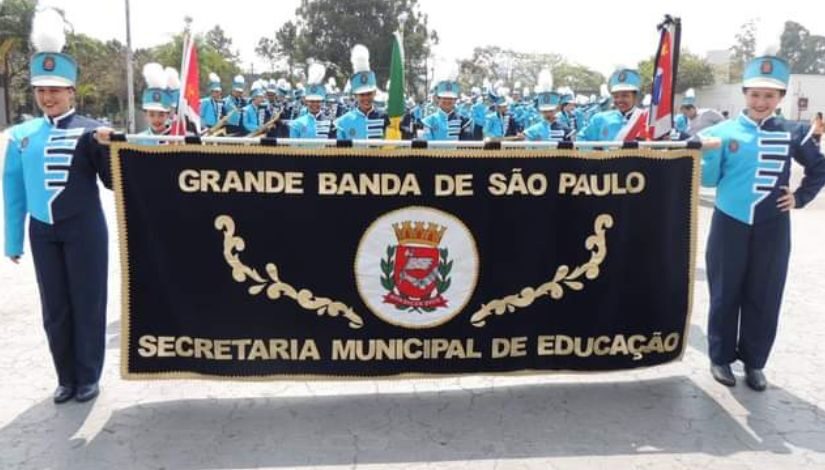 Foto de estudantes com roupa de desfile cívico, segurando uma faixa escrito: Grande Banda, Secretaria Municipal de Educação
