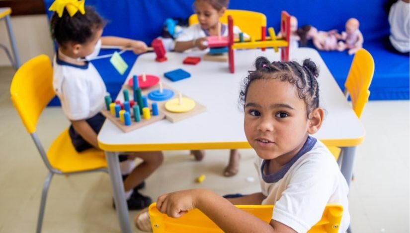 Criança está sentada olhando para a foto, ela está ao redor de uma mesa. Ao fundo pode-se ver outras duas meninas sentadas na mesma mesa brincando.