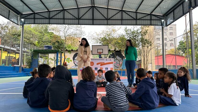 Estudantes sentados em roda observam educador conversando com eles dentro da quadra coberta