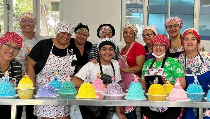 Fotografia de um grupo de estudantes do curso de confeitaria do Centro Municipal de Capacitação e Treinamento. Todos estão atrás de uma mesa e sobre ela o bolo de princesa que cada um produziu.