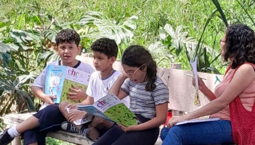 Fotografia mostra três crianças sentadas em um banco com livros na mão e a professora ao lado delas com um livro também. Ao fundo plantas verdes variadas.