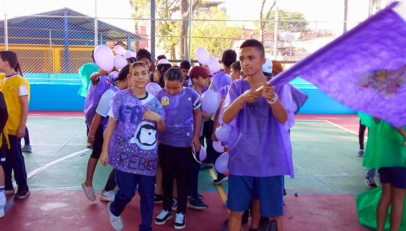 Estudantes estão de pé em fila, eles usam coletem feitos em tnt na cor roxa. O menino da frente segura uma bandeira roxa.