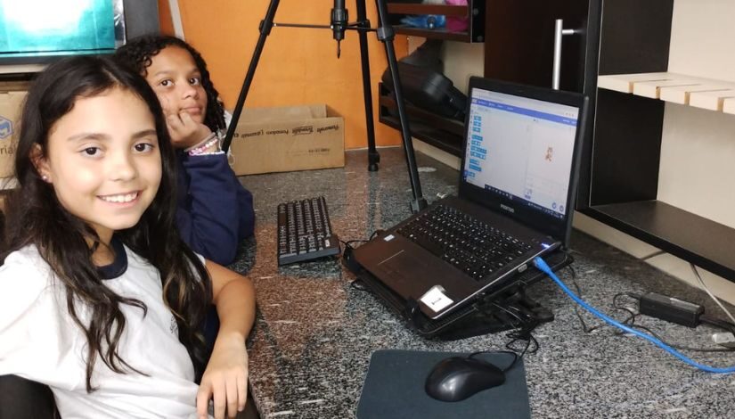 Duas meninas estão sentadas em frente a um notebook que está sobre uma mesa de mármore.