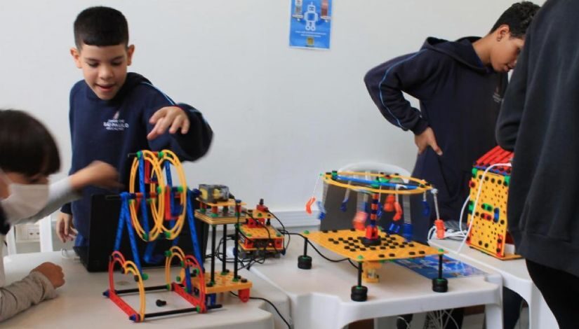 Fotografia mostra estudantes com uniforme azul marinho da prefeitura de SP. Eles manuseiam kits de robótica que estão sobre a sobre a mesa.