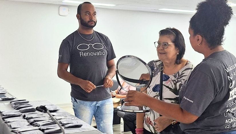 Fotografia mostra uma senhora com óculos, uma mulher segurando um espelho e um homem ao lado da senhora segurando uma caneta.