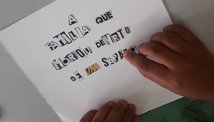Fotografia de uma folha de papel branca com letras recortadas onde se lê "A família que morou dentro de um sapato" e as mãos de uma criança.