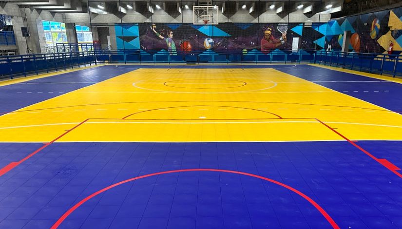 Fotografia de uma quadra poliesportiva com as cores azul, amarela e linhas vermelhas. Ao fundo nas paredes, há grafites de esportistas.