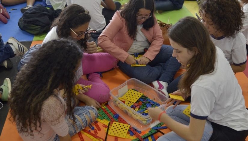 Estudantes sentadas em roda sobre um tapete de E.V.A. com peças coloridas do kit de robótica.