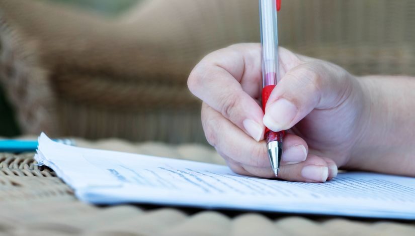 Fotografia mostra uma mão segurando uma caneta prata e vermelha sobre uma folha de papel com escritos desfocados.