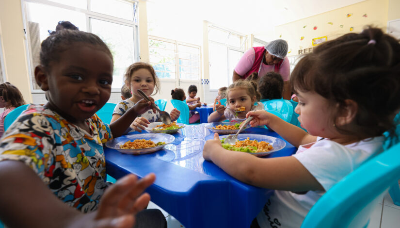 Fotografia de crianças pequenas se alimentando sentadas a mesa. 