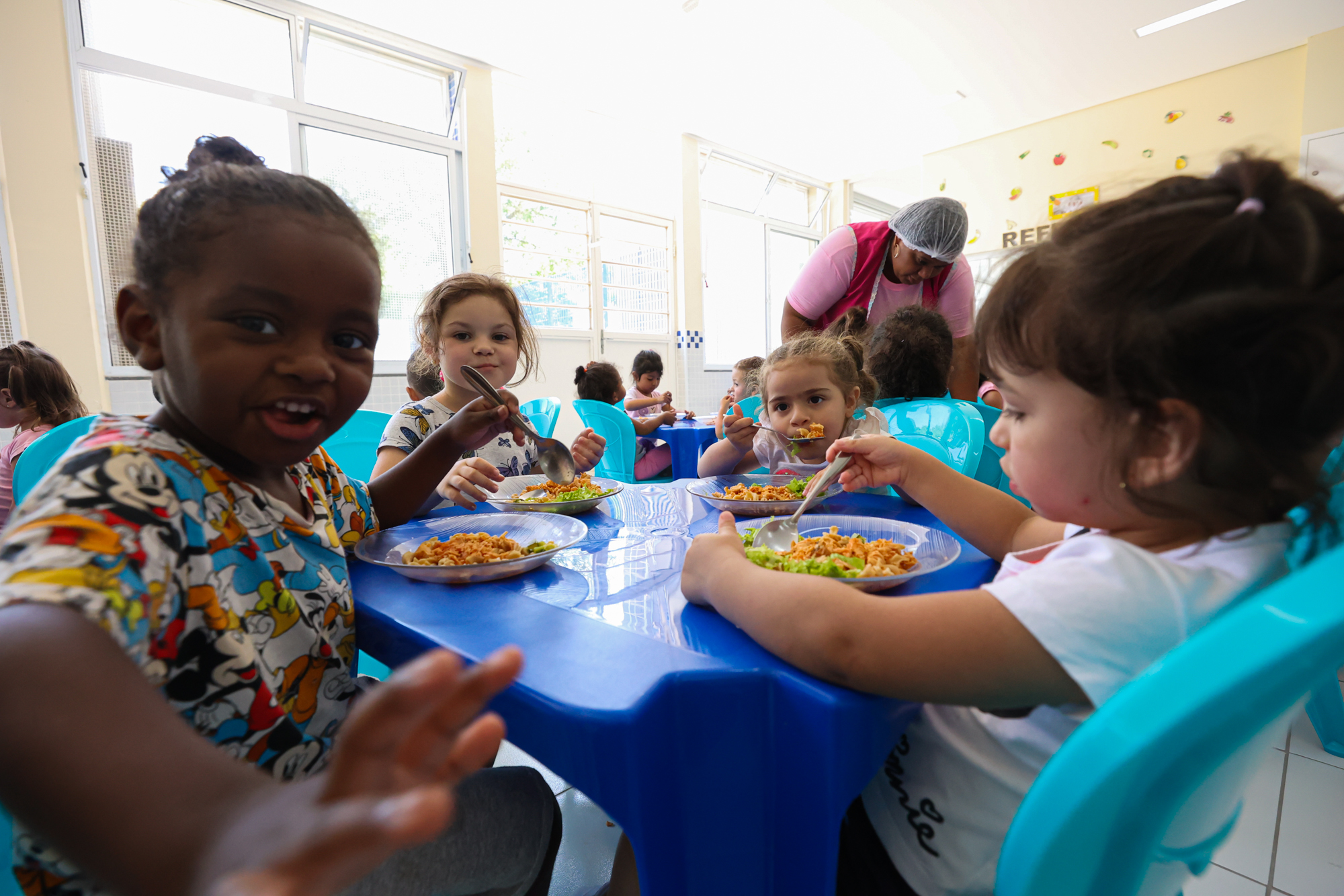 Fotografia de crianças pequenas se alimentando sentadas a mesa.