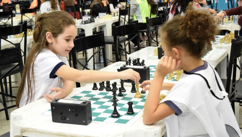 Fotografia mostra duas crianças sentadas ao redor de uma mesa, elas estão jogando xadrez.