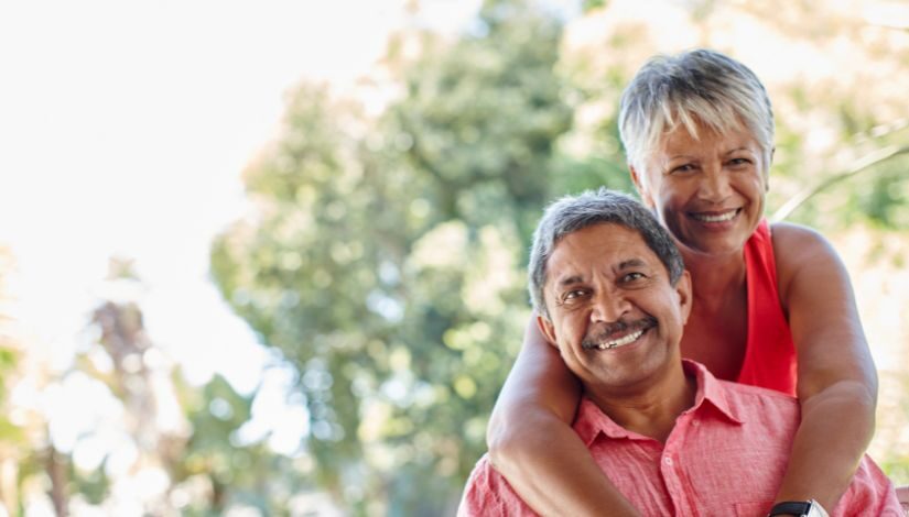 Imagem com árvores ao fundo e em primeiro plano uma mulher abraçando um homem, os dois estão sorrindo.