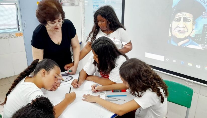 Fotografia mostra professora e estudantes em momento de atividade em sala de aula.