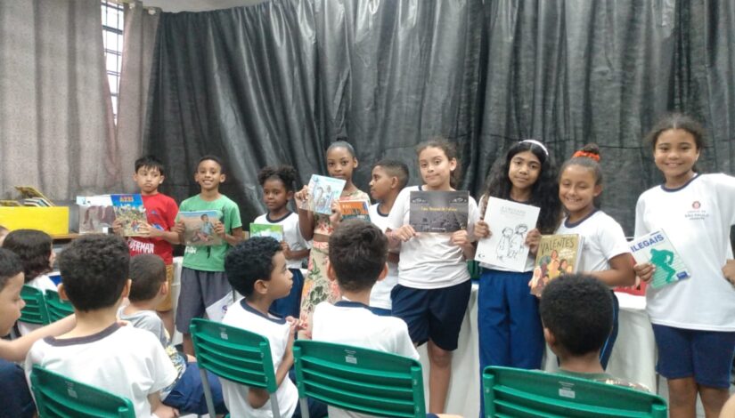 Fotografia de vários estudantes em pé. cada um deles segura um livros em suas mãos e mostram para os colegas que estão sentados a sua frente. 