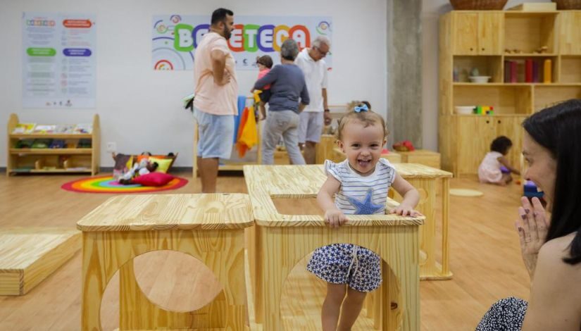 Uma criança sorri em um brinquedo e madeira, ao fundo um letreito colorido escrito "Bebeteca"