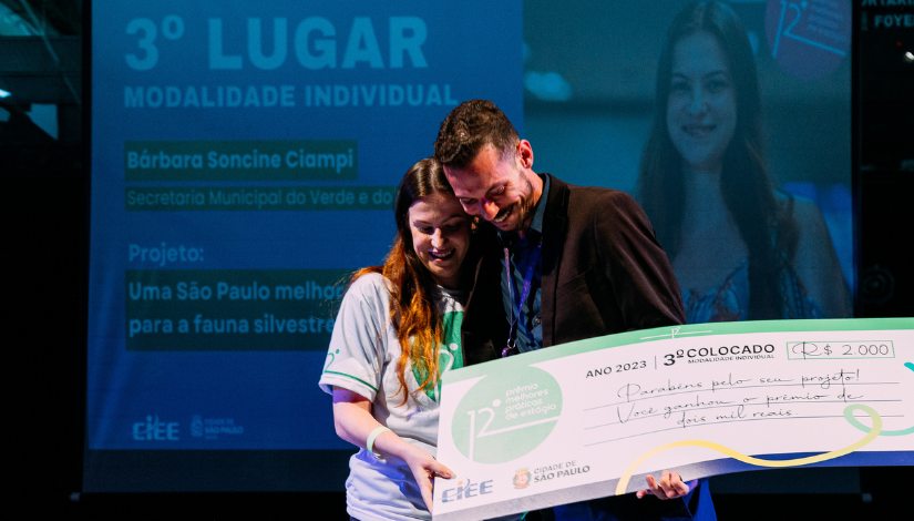 Fotografia mostra Bárbara Soncine Ciampi, à esquerda, comemorando o 3° lugar. Ela está segurando o cheque gigante do prêmio nas mãos. Ao fundo, um telão mostrando a foto dela e o nome do projeto
