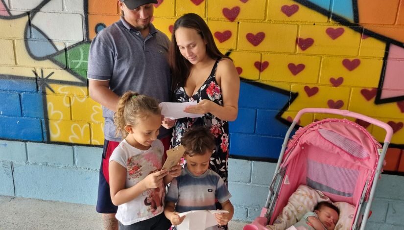 Fotografia de uma família em pé - há um homem, uma mulher, uma menina e um menino. Eles estão com cartas em suas mãos e olham para elas. No canto direito, há um carrinho de bebê rosa com uma bebê dentro. A parede ao fundo está com pintura de grafite colorida em cores amarela, vermelha e tons de azul.