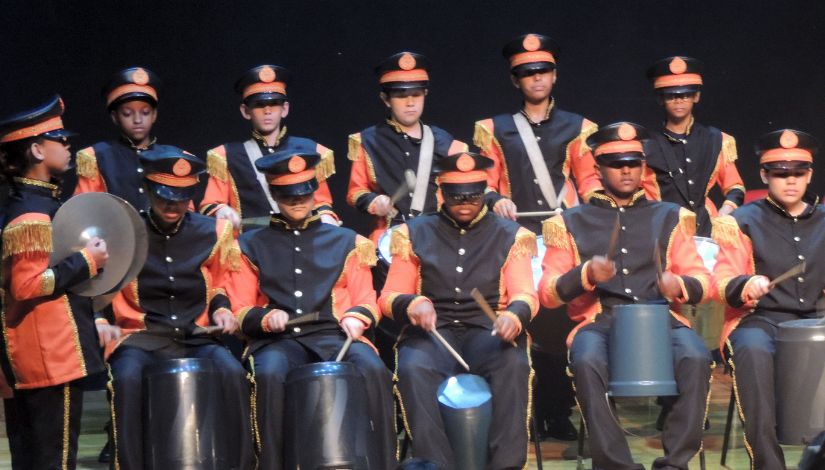 Fotografia mostra vários integrantes de uma fanfarra vestidos com uniforme azul marinho e laranja e seus instrumentos musicais.