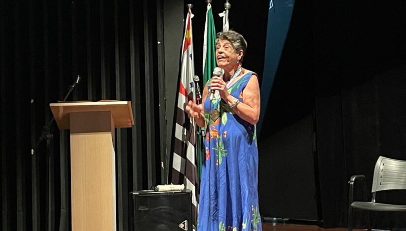 Fotografia mostra a socióloga Silvana Gimenes. Ela está em pé no palco, com um vestido azul estampado com flores e segurando o microfone. Ao fundo, aparece apenas a bandeira do estado de São Paulo.