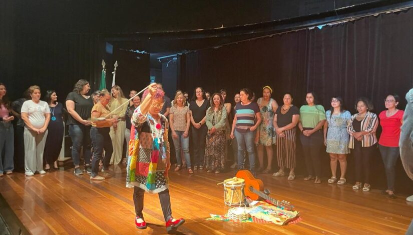 Fotografia mostra a professora Maria Maranhão ao centro de um palco com vários educadores em um semicírculo ao seu redor.