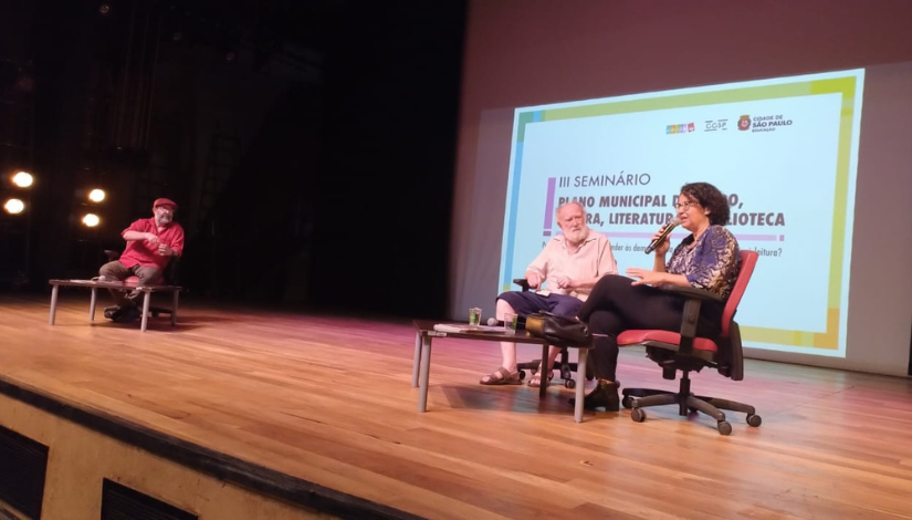 Fotografia mostra 3 pessoas em um palco de um teatro. Ao fundo percebe-se um telão com o texto III Seminário Plano Municipal do Livro, Leitura, Literatura e Biblioteca