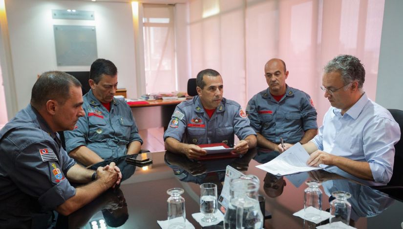 Fotografia mostra o secretário municipal de Educação, Fernando Padula, sentado à mesa com quatro bombeiros uniformizados. Ele está assinando um documento. 