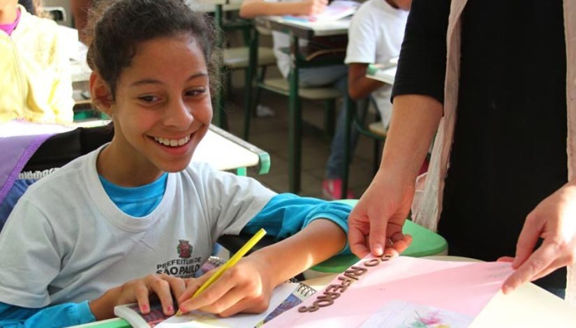 Fotografia de uma estudante sorrindo e segurando um lápis de cor amarelo sobre uma folha.