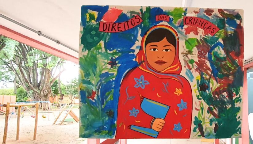 Fotografia mostra um tela com a pintura colorida da imagem da Malala escrito "Direito da Criança". Ao fundo, há um parque infantil.