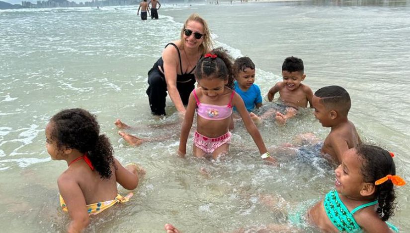 Crianças e uma mulher adulta dentro da água na praia. As crianças estão sentadas na parte rasa. 