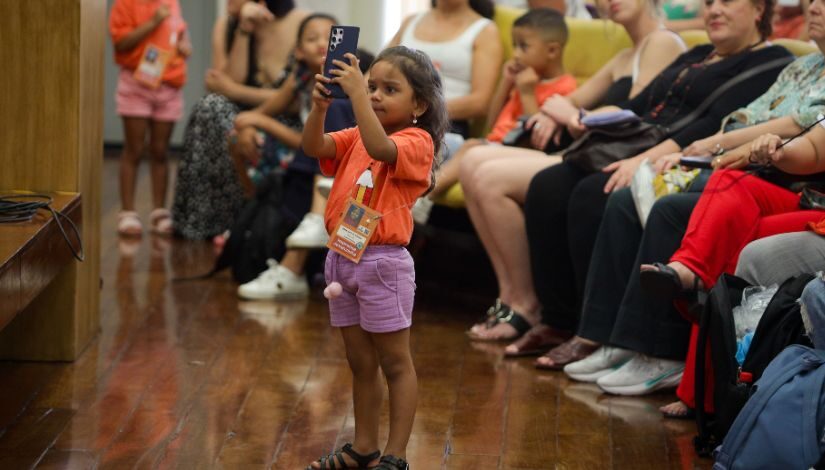 Uma menina pequena de camiseta laranja e shorts rosa com um celular na mão