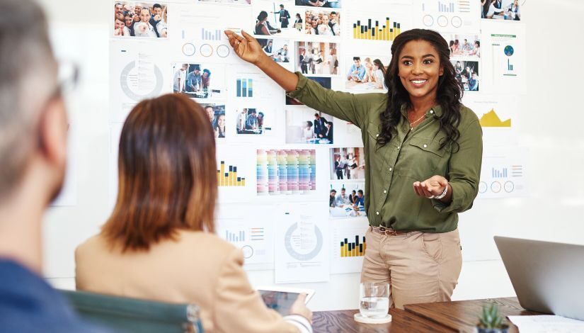 Fotografia de uma mulher negra com cabelos longos. Elá está em frente a um painel cheio de gráficos e outras imagens. Ela aponta com a mão direita para um dos gráficos e olha para duas pessoas que estão sentadas de costas para a câmera.