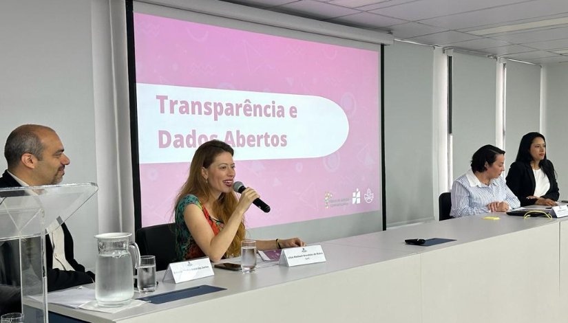 Uma mulher sentada a frente de uma mesa fala em um microfone, de fundo um telão escrito "Transparência e Dados Abertos
