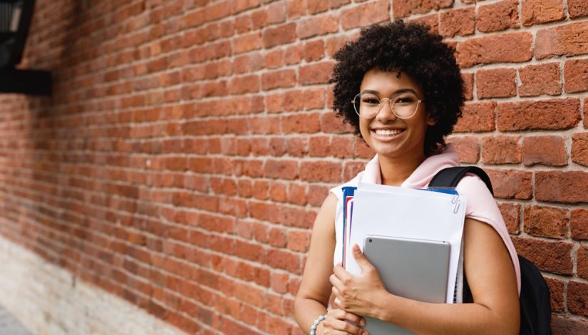 Fotografia de uma estudante negra. Ela está sorrindo, usa óculos, segura cadernos e tablet.