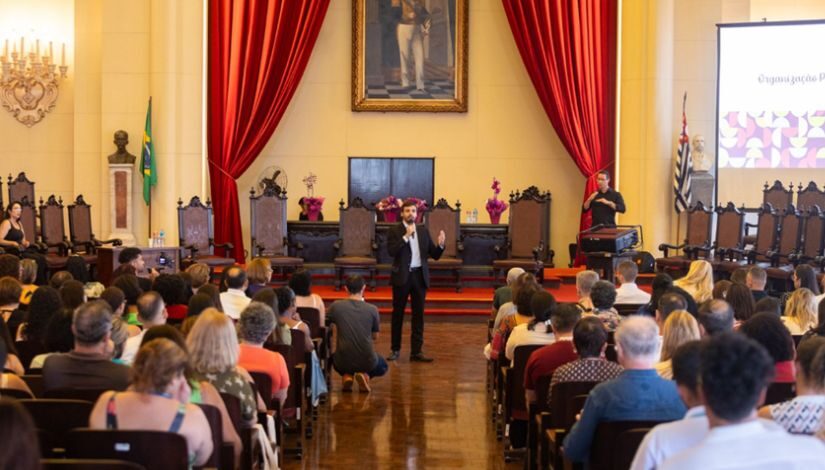 foto de um homem fazendo apresentação com pessoas sentadas em um auditório. ao fundo, uma cortina vermelha de veludo e um quadro, que compõe o auditório da faculdade de direito